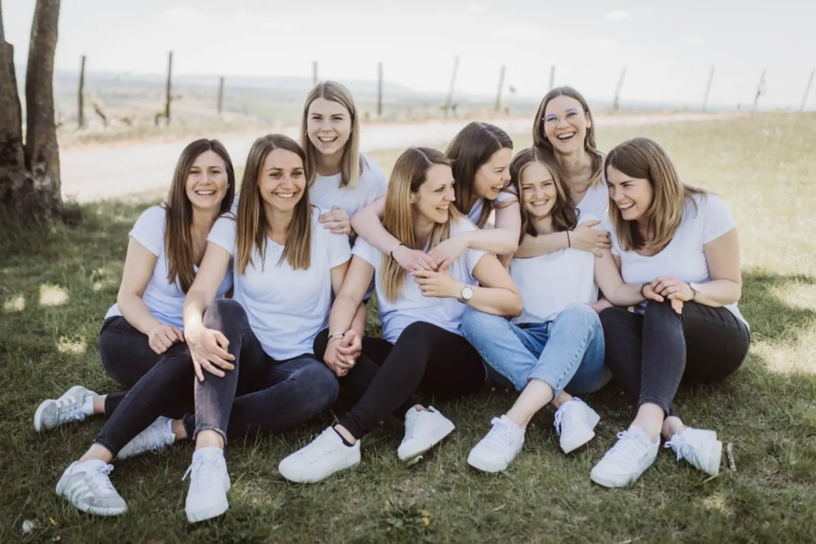 Gruppe von Frauen in weißen T-Shirts, die fröhlich im Freien sitzen und miteinander lachen.