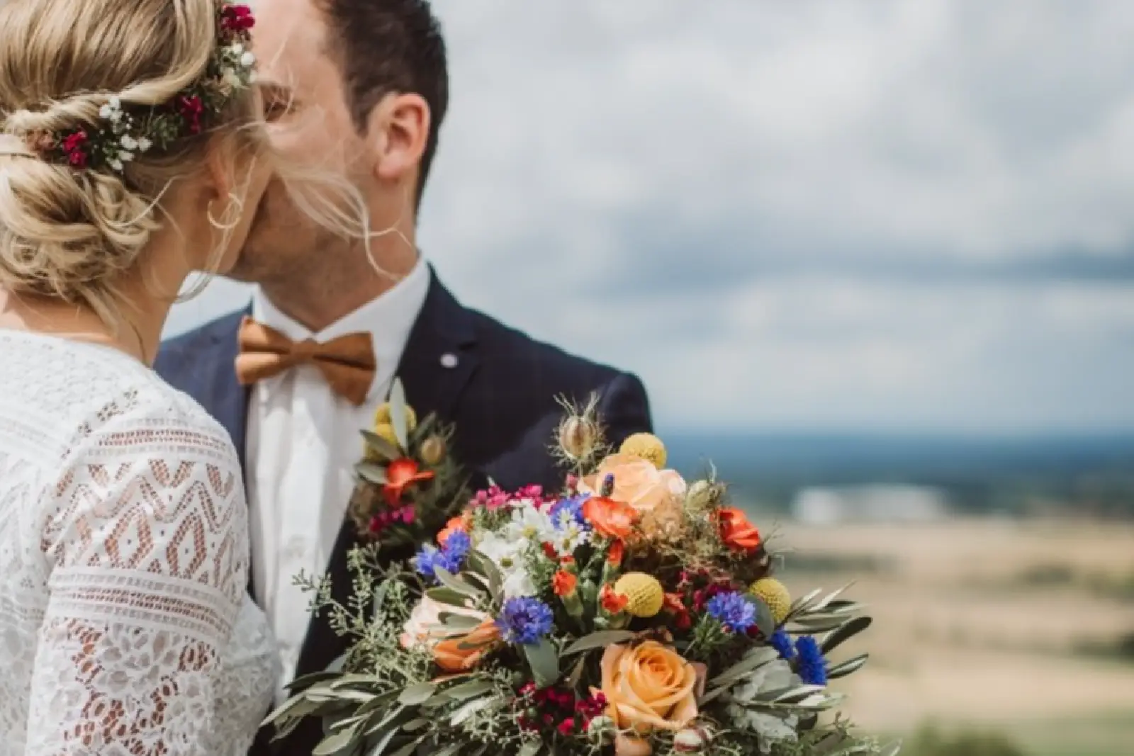 Das Brautpaar küsst sich, während die Braut einen bunten Blumenstrauß hält. Im Hintergrund ist eine landschaftliche Kulisse zu sehen.